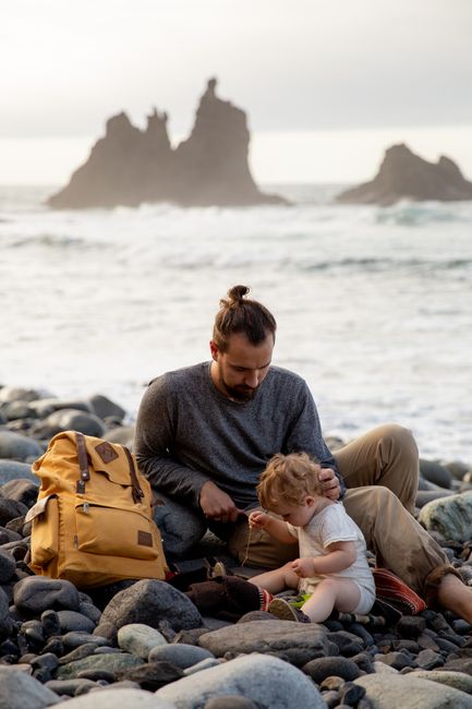 Tranquil man sitting near backpack with cute toddler on pebble coast near waving sea during vacation in soft daylight
