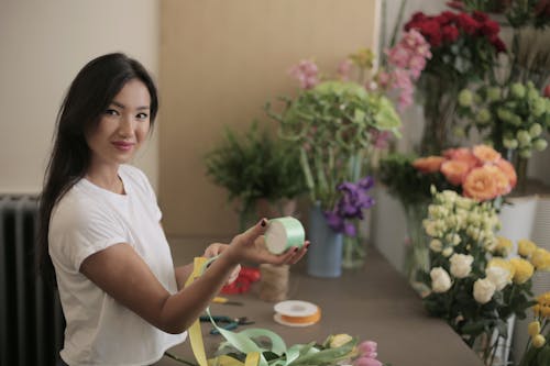 Young satisfied female florist working in flower store with colorful bands in rolls near counter with fresh bouquets while smiling and looking at camera