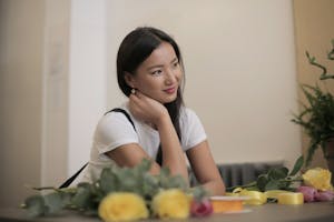Satisfied young Asian female florist with colorful silk bands in rolls for decoration of bouquets from fresh roses sitting at table and looking away with smile working in flower store