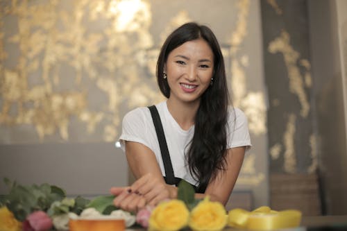 Positive ethnic female florist wearing apron smiling and looking at camera while standing near counter with fresh roses and silk tapes for composing bouquet inside flower store