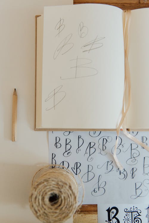 From above of white surface with minimalist composition of opened notebook with pencil and sheet of paper with printed letters together with other decorative elements illustrating handwriting practice