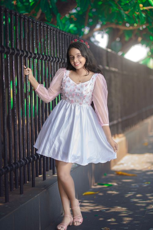 Free A young woman in a white dress with pink sleeves poses cheerfully by a fence outdoors. Stock Photo