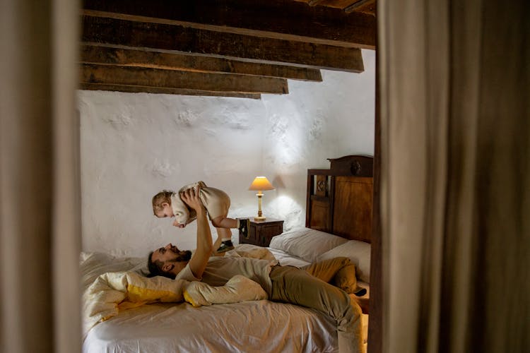 Happy Bearded Man With Toddler Lying On Bed In Rural Interior House