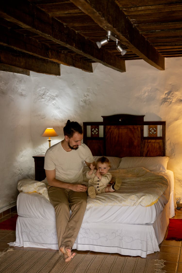 Happy Bearded Man With Toddler Sitting On Bed In Rural Interior House