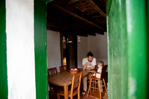 View through entrance of adorable little kid spending time together with bearded father in casual outfit inside aged room near wooden desk with wooden chairs during daytime