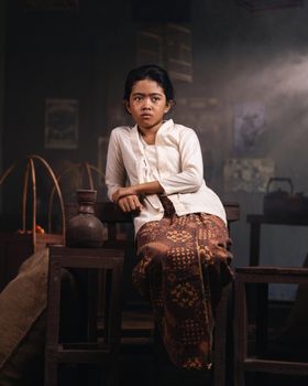 A young girl in traditional Indonesian clothing sits indoors, bathed in soft light.