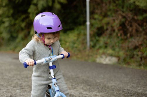 Cute toddler child wearing protective helmet and warm bodysuit playing with bicycle on road near green plants on rural street