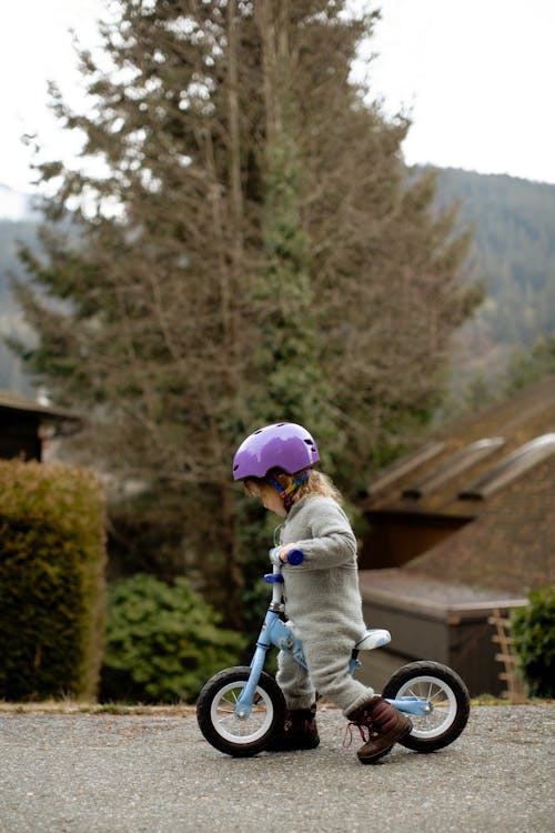 Side view of cute little girl in protective helmet and warm clothes riding balance bike along asphalt path on autumn street in countryside