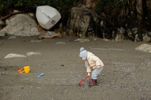 Little kid in warm clothes playing with toy shovel on sandy beach