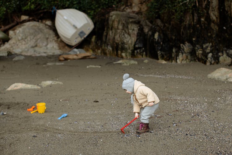 Little Kid In Warm Clothes Playing With Toy Shovel On Sandy Beach