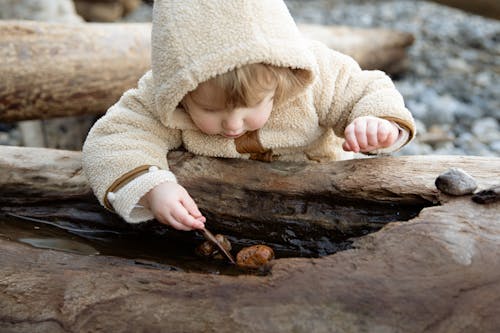 Cute little girl in warm coat playing with small pebbles on wet log while resting on shore in autumn day