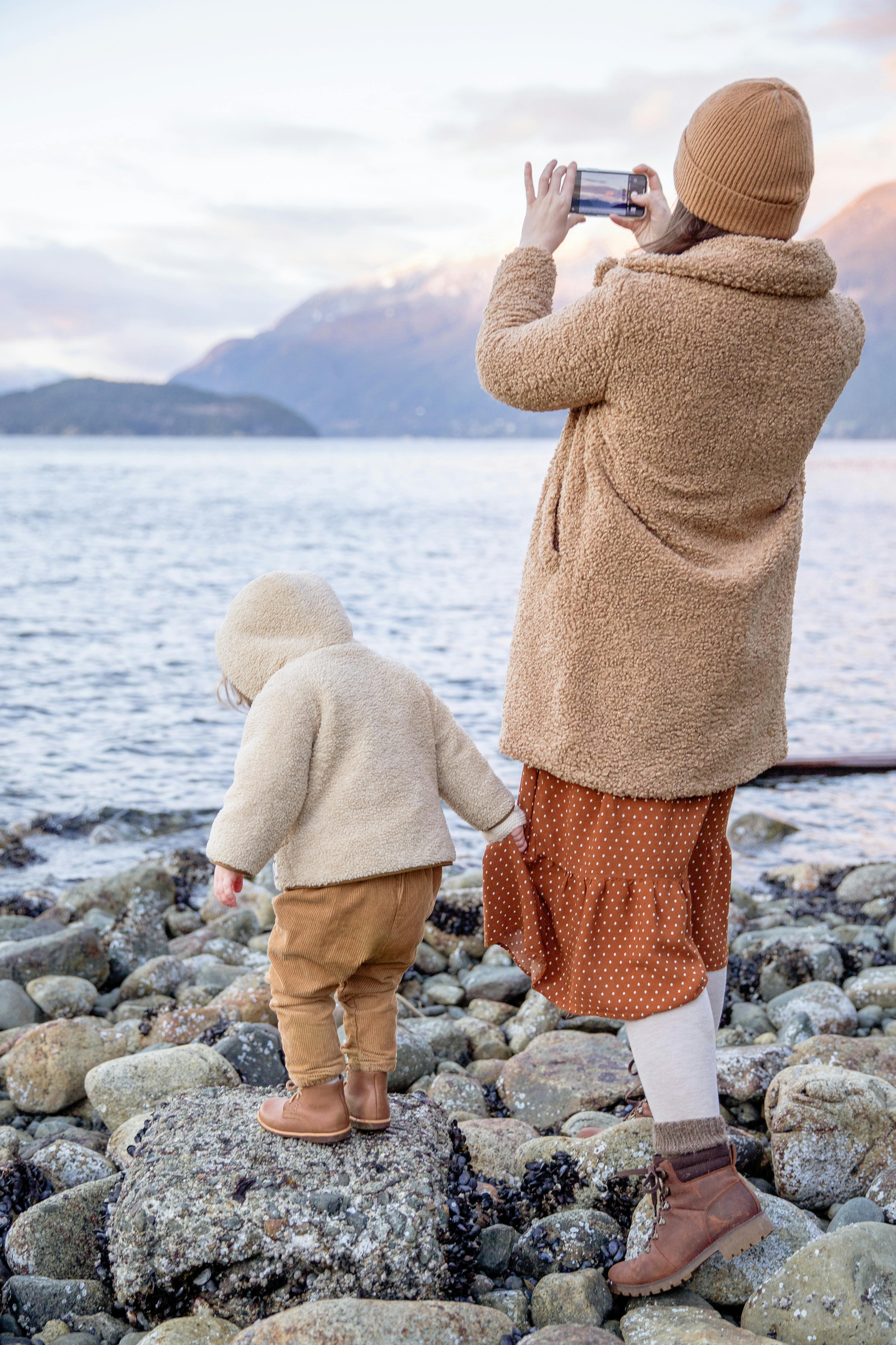 mother and child in warm clothes standing on rocky seashore near water and taking photo on smartphone