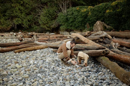 Free Father and little child wearing warm clothes playing with stones on rocky ground near logs and forest during weekend trip Stock Photo