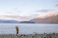 一位母亲抱着孩子站在平静水边的石岸上，身后照片已面目全非，他们在旅途中欣赏着如画的山景