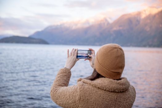 Back view of unrecognizable female tourist in warm coat and hat taking photo on smartphone while standing on coast near water and enjoying seascape during trip