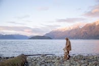 母亲和小孩穿着休闲保暖外套，手牵着手，沿着石岸在平静的水面旁散步的侧面图