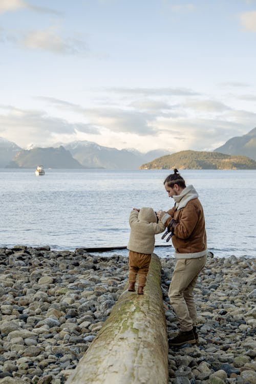 Bearded father in casual warm clothes helping little kid to balance on big log on stony coast near calm water during weekend in autumn