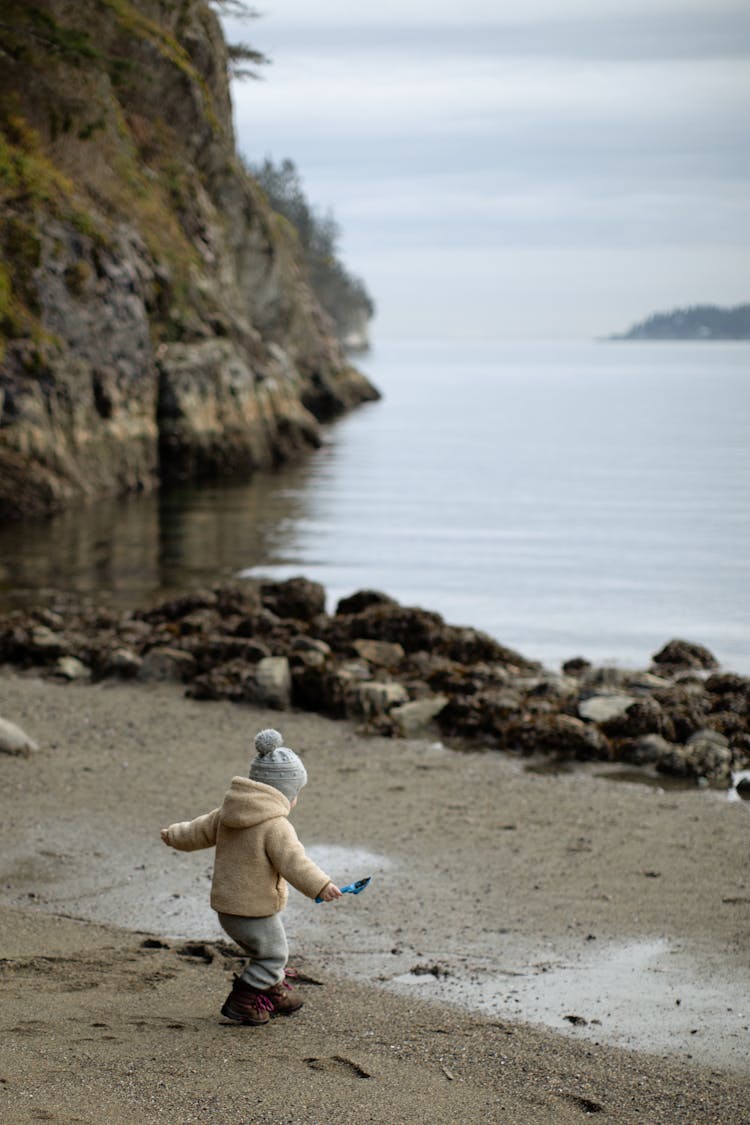 Unrecognizable Little Child Playing On Sandy Shore Near Mountain