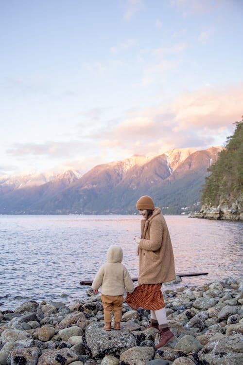 Back view unrecognizable preschool boy in warm outerwear holding skirt of mother in warm clothes and hat while traveling on calm lake in autumn