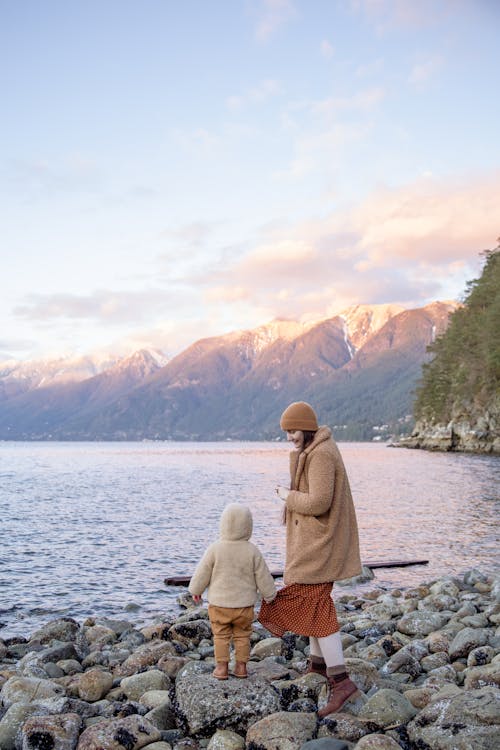 Free Back view unrecognizable preschool boy in warm outerwear holding skirt of mother in warm clothes and hat while traveling on calm lake in autumn Stock Photo