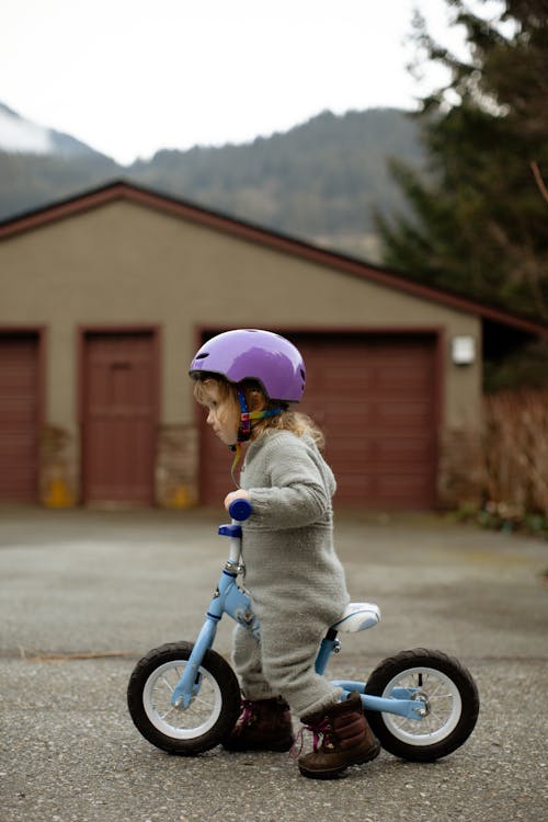 Side view of adorable little girl in protective helmet and casual warm clothes riding balance bicycle against garage building down street