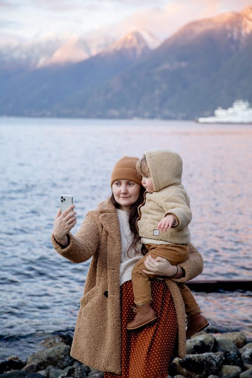 Calm woman carrying adorable child on hands and taking selfie on smartphone near river and mountains