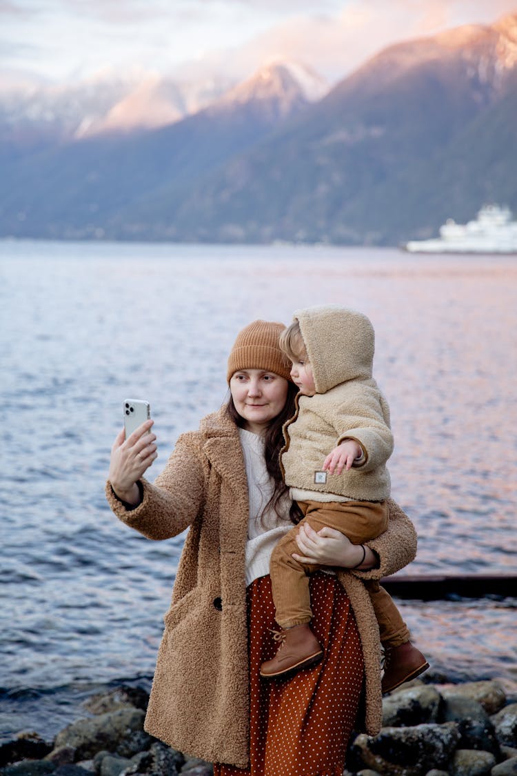 Calm Woman Carrying Adorable Child On Hands And Taking Selfie On Smartphone Near River And Mountains