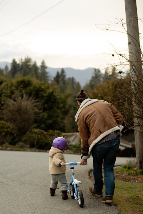 Free Back view of unrecognizable man and toddler wearing warm wear and walking on street with balance bicycle near green trees in countryside Stock Photo