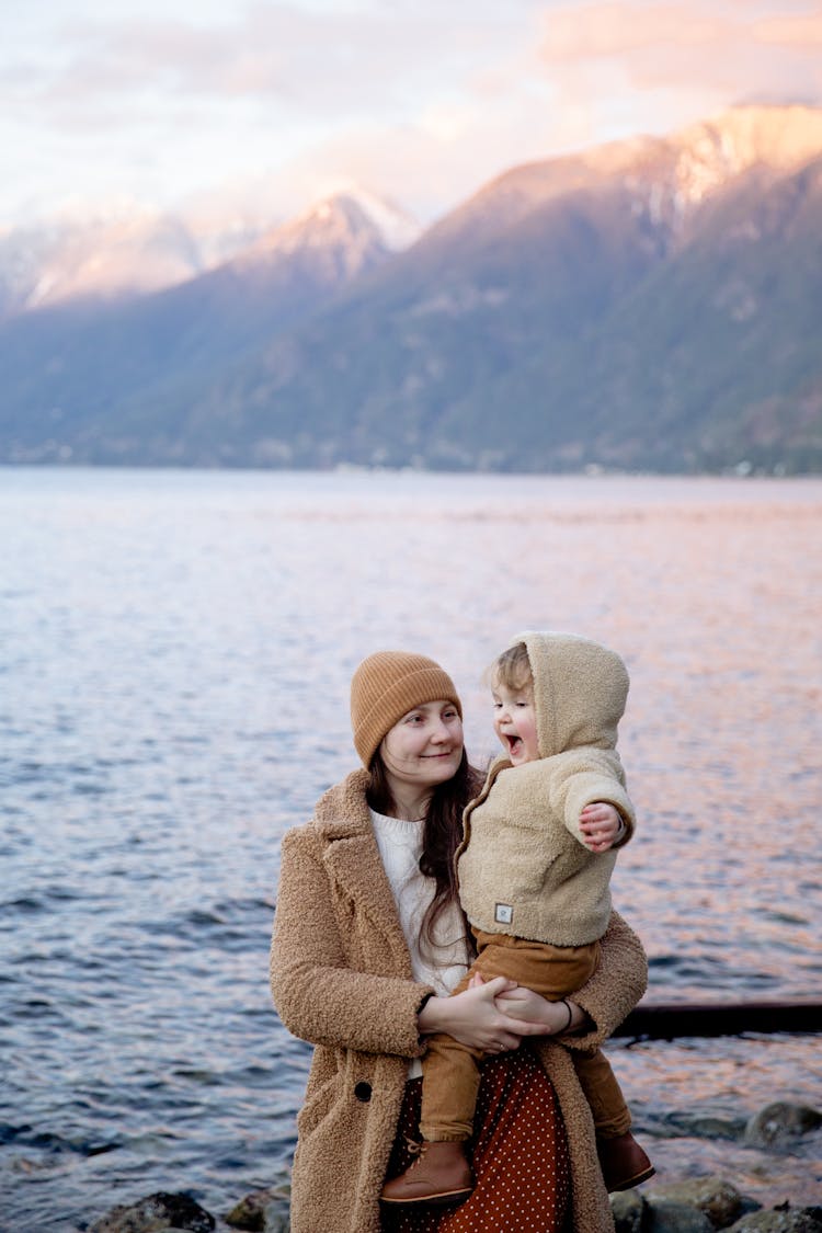 Positive Mother Holding Cute Baby During Vacation Near Seashore In Winter