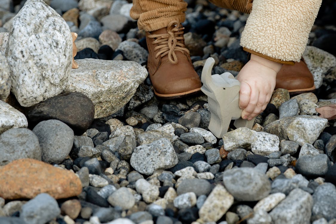 Unrecognizable little child in warm clothes and brown boots playing with stone toy of elephant between stones on seashore in winter