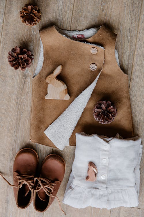 Top view of children boots leather brown vest and linen wear placed on floor decorated with cones and wooden rabbits