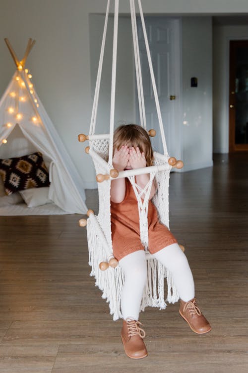 Free Little kid covering face with hands while chilling in hanging chair Stock Photo