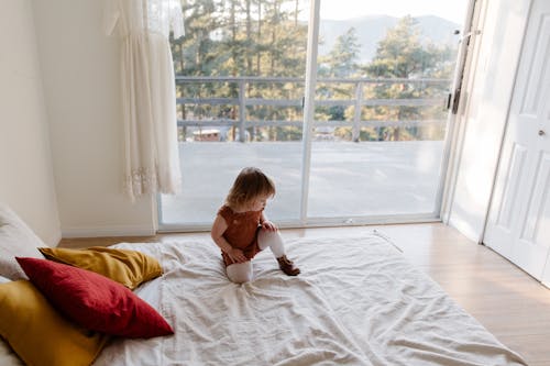 From above of adorable little child in casual clothes playing on bed against wide window while spending time at home