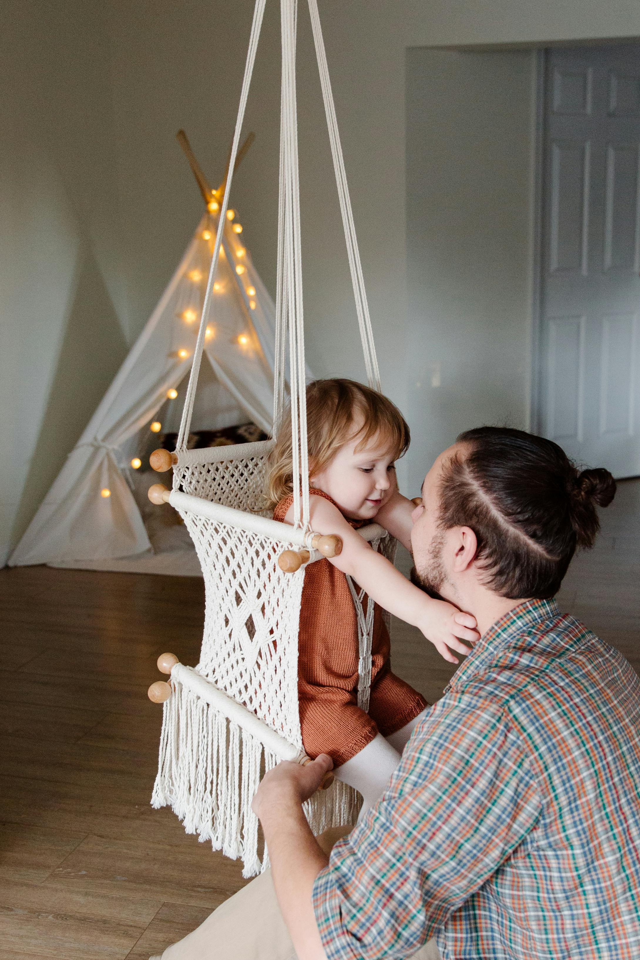 Happy little girl sitting on woven swing and holding out hands to father while spending time together in cozy room and looking at each other