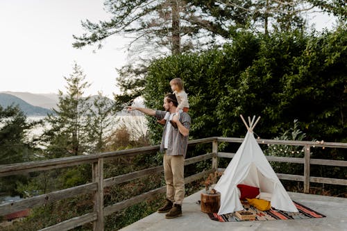 Side view of little child sitting on shoulders of father while man pointing to mountain while standing on wooden terrace with wigwam and picturesque view