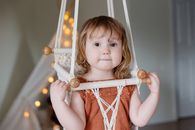 室内，可爱的幼儿坐在织物秋千上，周围环绕着串灯，营造出舒适的氛围