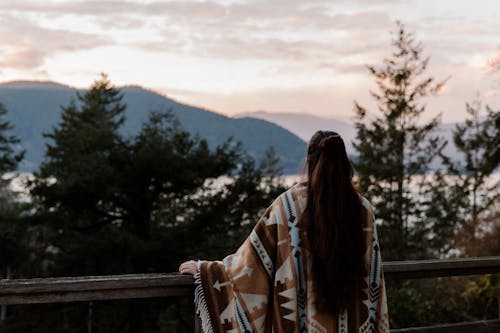 Back view of unrecognizable female covered warm blanket with ethnic prints standing on viewpoint and observing landscape