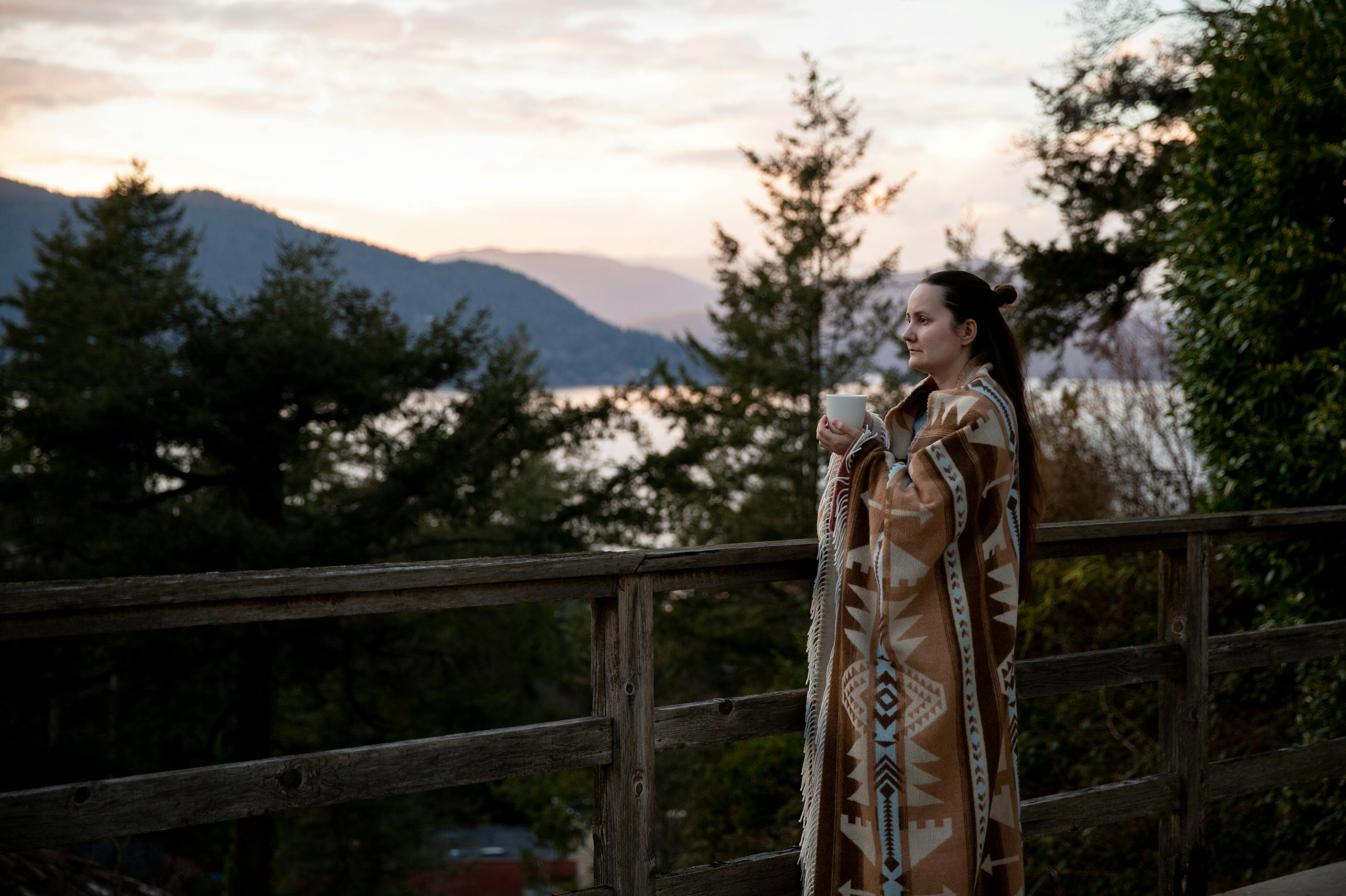 A woman wrapped in a blanket enjoys a warm cup of coffee on a wooden deck overlooking a peaceful, scenic sunset.