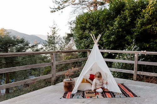 Adorable little kid sitting on rug in wigwam located on terrace with wooden border and scenic view