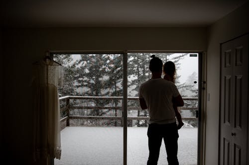 Back view of anonymous man holding kid while standing opposite of window in room and looking at snowy day