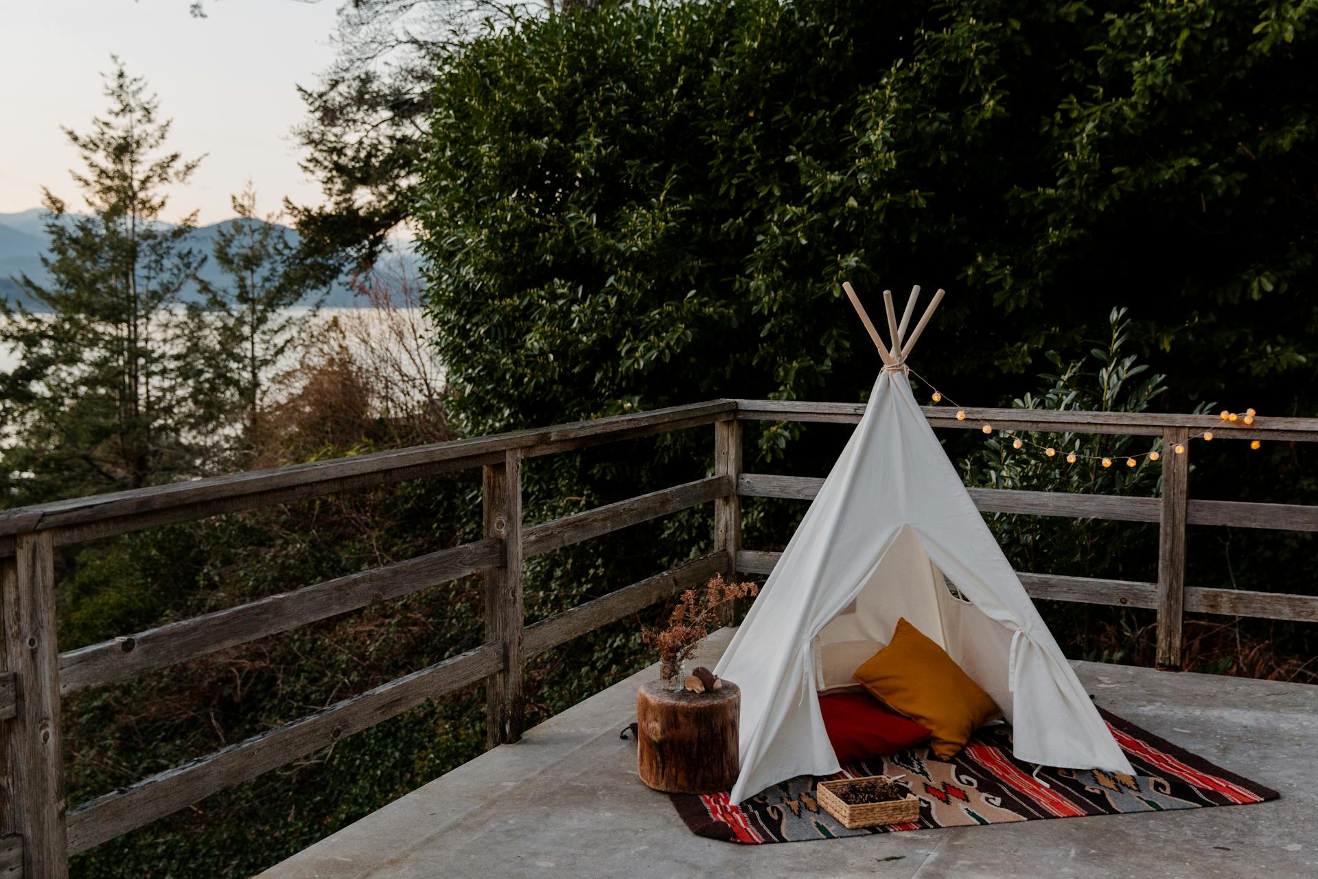 Boho styled tent with rug and pillows located on terrace with wooden fence with picturesque view to sea forest and mountains