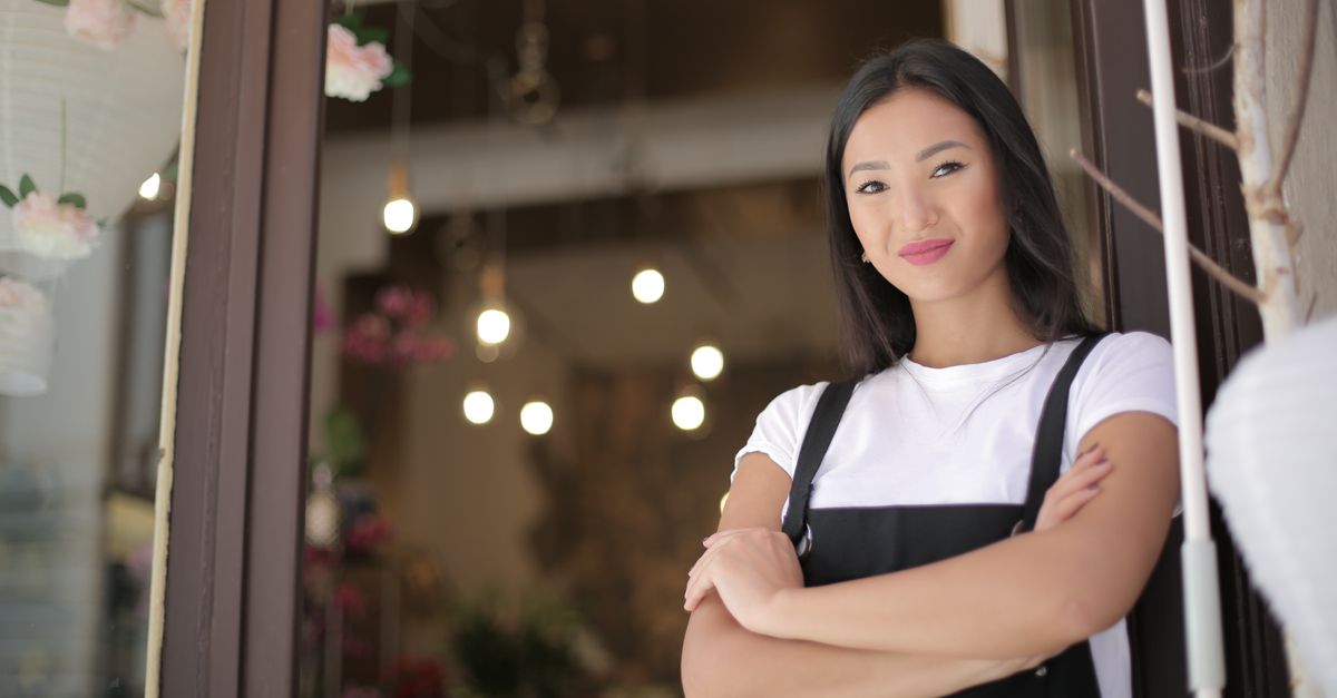 From below of young confident Asian female standing with crossed arms at entrance of floral shop and smiling at camera