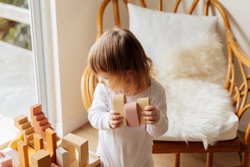 From above of cute little child in white wear holding wood blocks for building while standing near wicker armchair