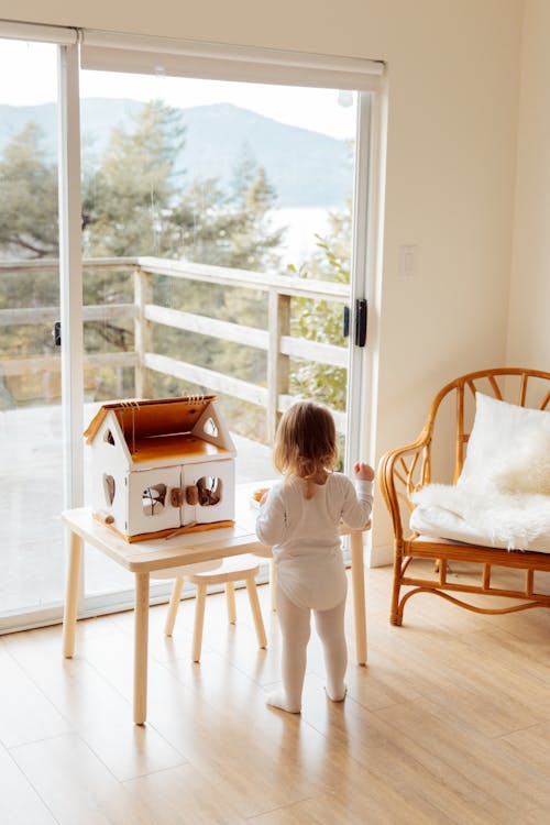 Cute little girl playing with toy house at table near window