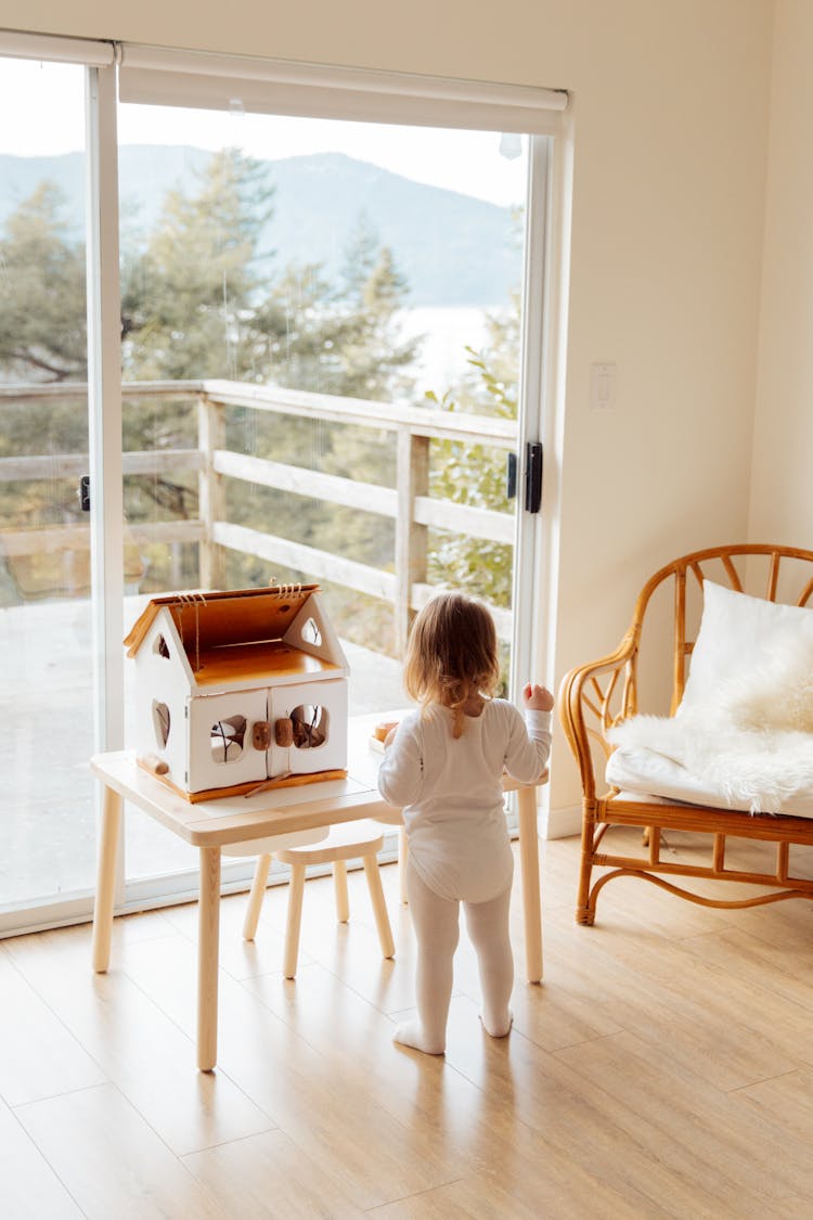 Cute Little Girl Playing With Toy House At Table Near Window