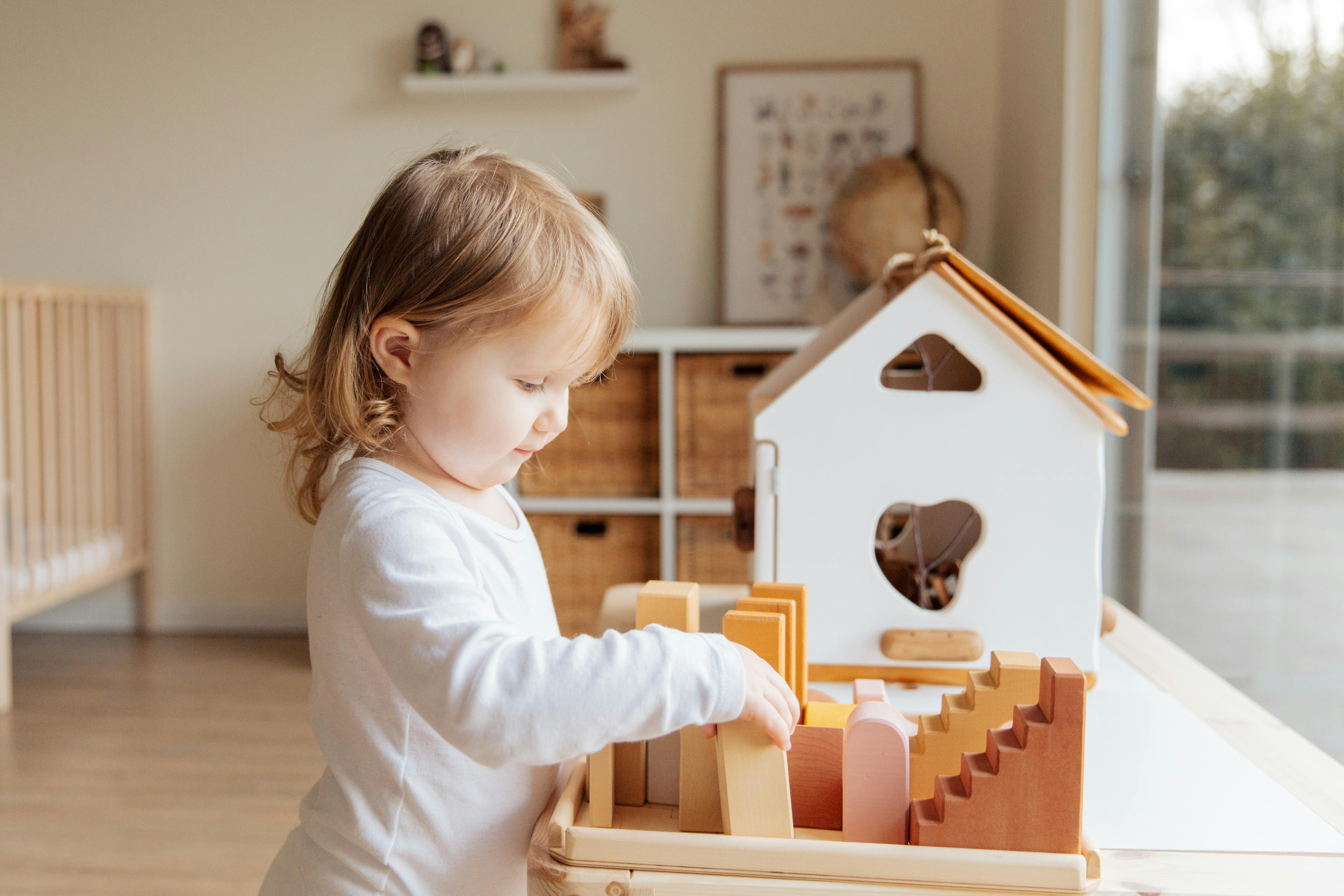 cute little girl playing with wooden blocks at table near window at home