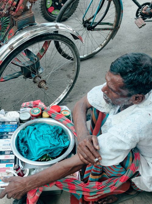 Free An adult street vendor in Dhaka offers cigarettes and traditional betel leaves beside a colorful rickshaw. Stock Photo