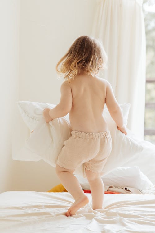 Back view of anonymous little girl in casual clothes playing with pillows on comfortable bed in light room at home