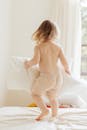 Back view of anonymous little girl in casual clothes playing with pillows on comfortable bed in light room at home