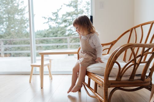 Side view of cute little girl in casual dress sitting on edge of comfortable armchair near table in light living room at home